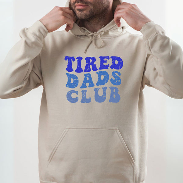 A man wearing a beige hoodie with the text "TIRED DAD'S CLUB" printed on it.