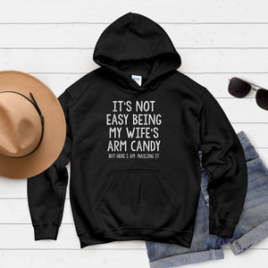 A black hoodie with white text that reads "IT'S NOT EASY BEING MY WIFE'S ARM CANDY BUT HERE I AM MAILING IT" is displayed on a white background.
