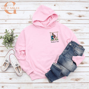 A pink hoodie with a picture of a couple on the front, a pair of blue jeans, and a pair of white sneakers are laid out on a white wooden surface.