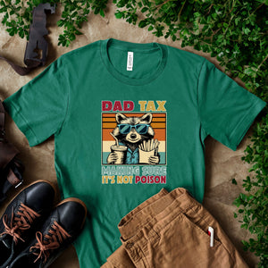 a green t-shirt with a graphic design of a raccoon wearing sunglasses and holding a drink, accompanied by the text "DAD TAX MAKING SURE IT'S NOT POISON". The t-shirt is laid out on a surface with some shoes and a pair of pants, and there are some plants in the background.
