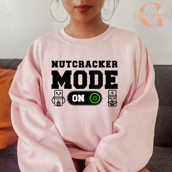 A woman wearing a pink sweatshirt with the text "NUTCRACKER MODE ON" printed on it.