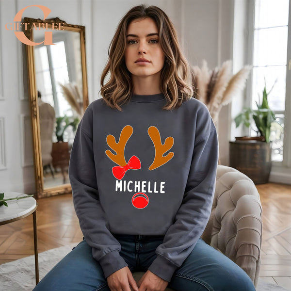 A woman wearing a gray sweatshirt with a reindeer and the name "Michele" printed on it, sitting on a chair in a room with a mirror and potted plants.