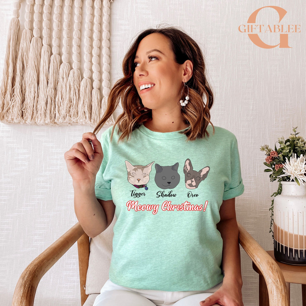 A woman wearing a light green t-shirt with a graphic of three cat faces and the text "Meowy Christmas!" is sitting in a chair.