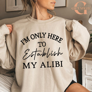 A woman wearing a beige sweatshirt with the text "I'M ONLY HERE TO ESTABLISH MY ALIBI" printed on it.