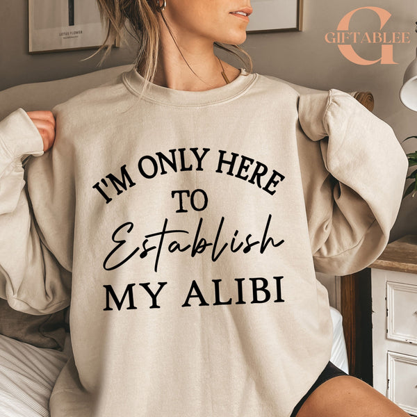 A woman wearing a beige sweatshirt with the text "I'M ONLY HERE TO ESTABLISH MY ALIBI" printed on it.