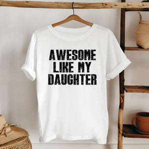 A white t-shirt with the text "AWESOME LIKE MY DAUGHTER" printed on it, hanging on a wooden hanger.
