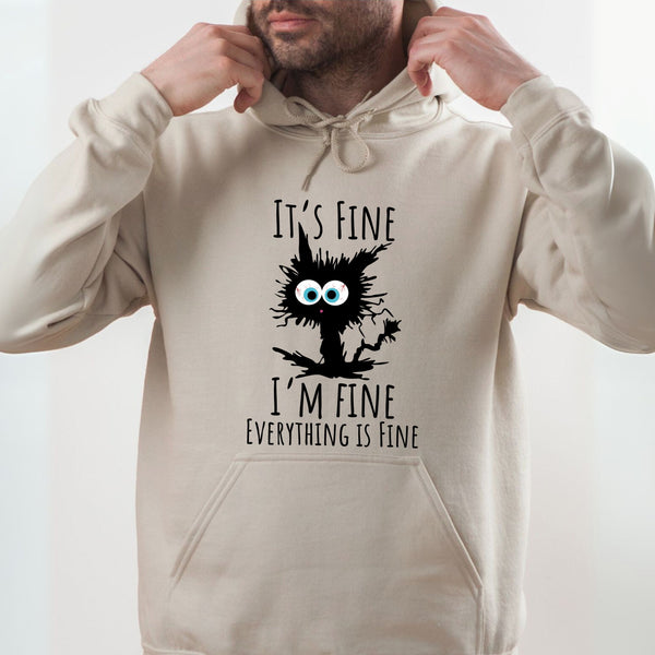 A man wearing a beige hoodie with a graphic design featuring a cat with a message that reads "It's fine I'm fine everything is fine".