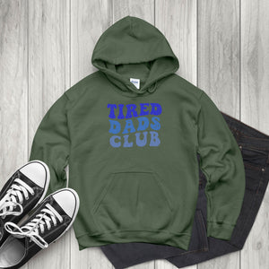 A green hoodie with the text "TIRED DAD'S CLUB" printed on it, along with a pair of black and white sneakers, is laid out on a wooden surface.