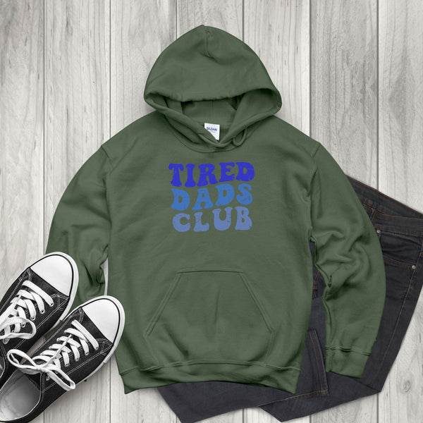 A green hoodie with the text "TIRED DAD'S CLUB" printed on it, along with a pair of black and white sneakers, is laid out on a wooden surface.