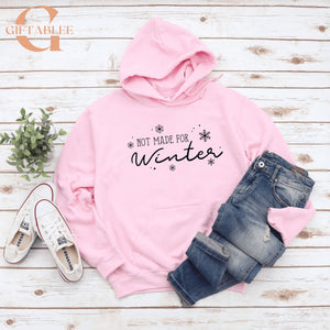 A pink hoodie with the text "NOT MADE FOR WINTER" printed on it, along with snowflakes, is displayed on a wooden surface alongside a pair of jeans and white sneakers.