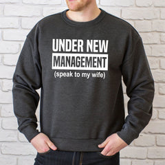 A man wearing a grey sweatshirt with the text "Under New Management (speak to my wife)" printed on it.