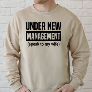 A man wearing a beige sweater with the text "Under New Management (speak to my wife)" printed on it.