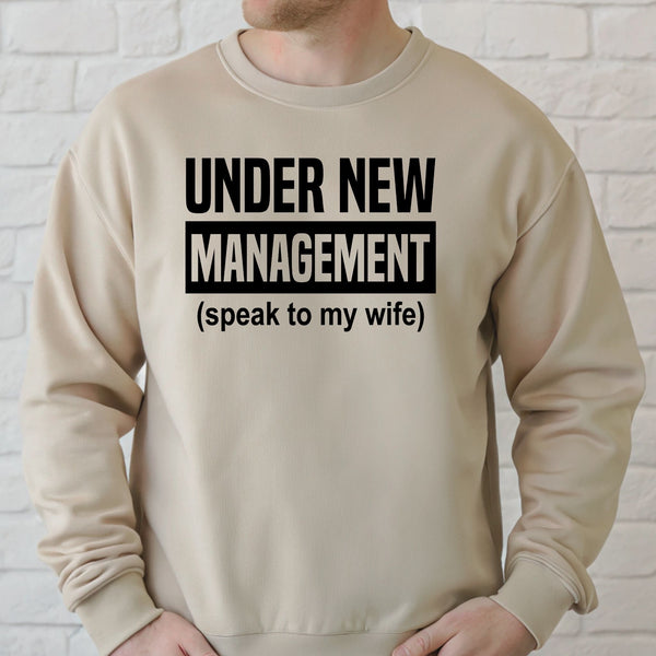 A man wearing a beige sweater with the text "Under New Management (speak to my wife)" printed on it.