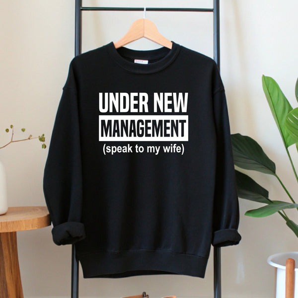 A black sweatshirt with white text that reads "UNDER NEW MANAGEMENT [speak to my wife]" hanging on a wooden hanger.