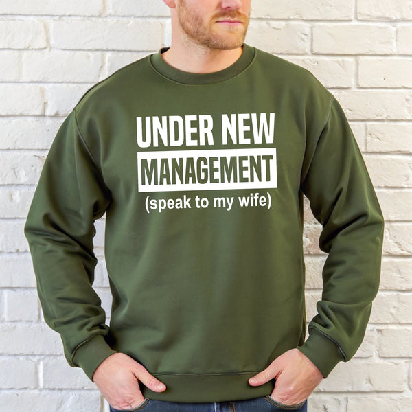 A man wearing a green sweatshirt with the text "UNDER NEW MANAGEMENT (speak to my wife)" printed on it.