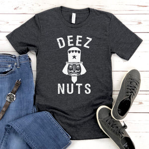 a heather dark gray t-shirt with the text "DEEZ NUTS" printed on it, along with a nutcracker graphic. The t-shirt is laid out on a wooden surface, accompanied by a pair of blue jeans, a watch, and a pair of sneakers.