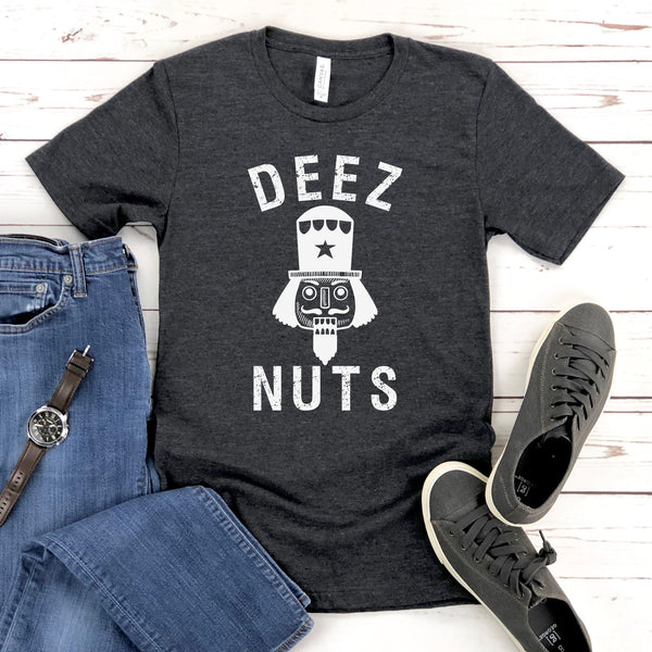 a heather dark gray t-shirt with the text "DEEZ NUTS" printed on it, along with a nutcracker graphic. The t-shirt is laid out on a wooden surface, accompanied by a pair of blue jeans, a watch, and a pair of sneakers.