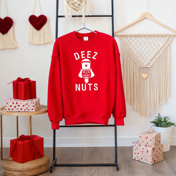 A red sweatshirt with the text "DEEZ NUTS" printed on it, hanging on a black hanger. The sweatshirt is displayed in front of a Christmas-themed background, which includes a macrame wall hanging, red gift boxes, and a potted plant.