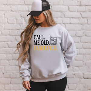 A woman wearing a gray sweatshirt with the text "Call me old. Fashioned" printed on it, standing in front of a white brick wall.