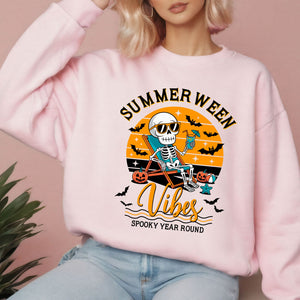 A woman wearing a pink sweatshirt with a graphic design featuring a skeleton in a skeleton chair, holding a drink, and the text "Summer Ween Vibes Spooky Year Round".