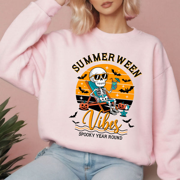 A woman wearing a pink sweatshirt with a graphic design featuring a skeleton in a skeleton chair, holding a drink, and the text "Summer Ween Vibes Spooky Year Round".