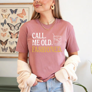 A woman wearing a pink t-shirt with the text "Call me old. Fashioned" printed on it, standing in front of a butterfly-themed wall art.