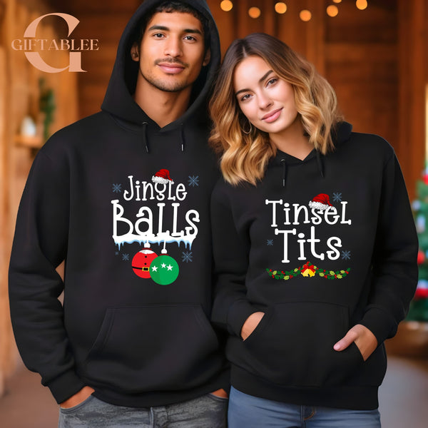 A man and a woman wearing matching black hooded sweatshirts with Christmas-themed text printed on them.