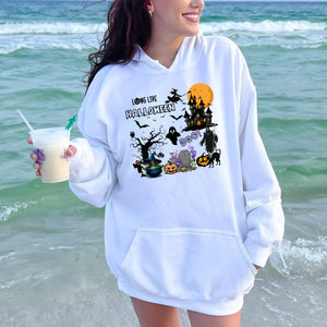 A woman wearing a white hoodie with a Halloween-themed graphic and holding a drink on a beach.