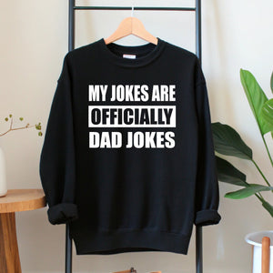 A black sweatshirt with white text that reads "MY JOKES ARE OFFICIALLY DAD JOKES" hanging on a wooden hanger.