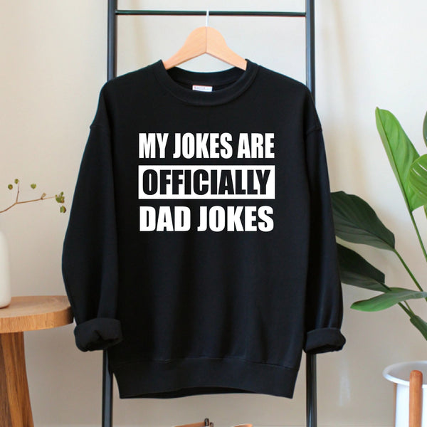 A black sweatshirt with white text that reads "MY JOKES ARE OFFICIALLY DAD JOKES" hanging on a wooden hanger.