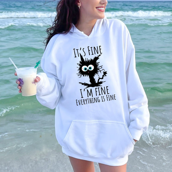 A woman wearing a white hoodie with a graphic design of a cat and the text "It's fine I'm fine everything is fine" is standing near the ocean.