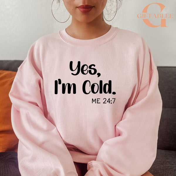 A woman wearing a pink sweatshirt with the text "Yes, I'm Cold. ME 24:7" printed on it.