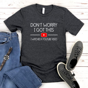 a gray t-shirt with the text "DON'T WORRY I GOTT THIS I WITNESSED A YOUTUBE VIDEO" printed on it, along with a red play button symbol. The t-shirt is laid out on a wooden surface, accompanied by a pair of blue jeans and a pair of gray sneakers.