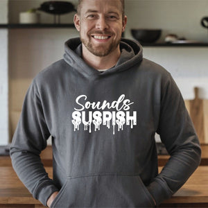 A man wearing a grey hoodie with the text "Sounds Suspicious" printed on it, smiling at the camera.