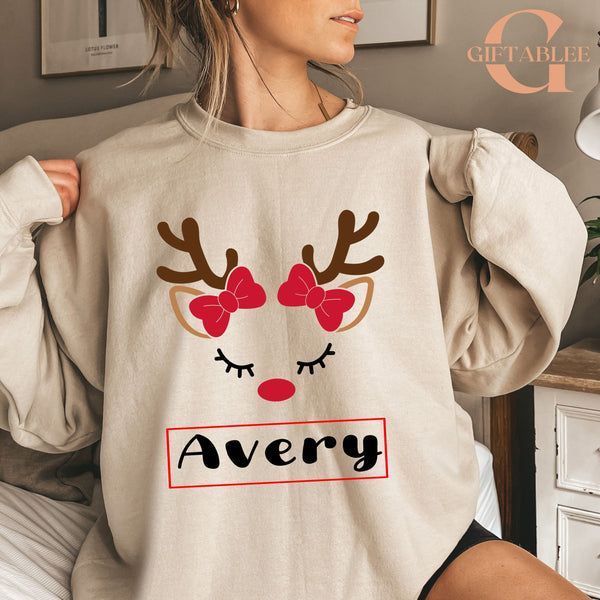 A woman wearing a beige sweater with a reindeer face design and the name "Avery" printed on it.