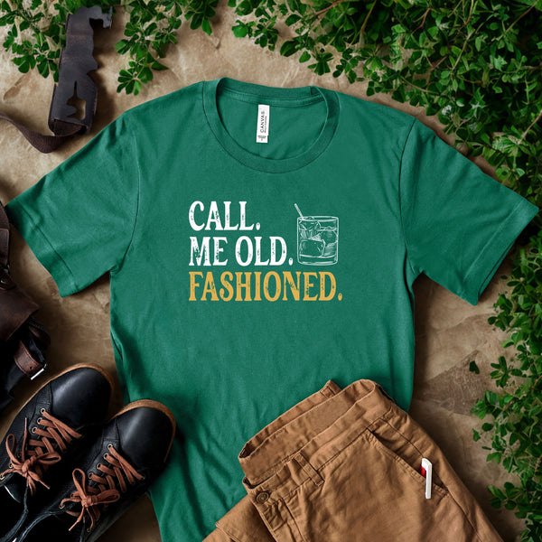 a green t-shirt with the text "Call me old, Fashioned." printed on it, along with a graphic of a cocktail glass. The t-shirt is laid out on a surface with some plants and a pair of shoes visible in the background.
