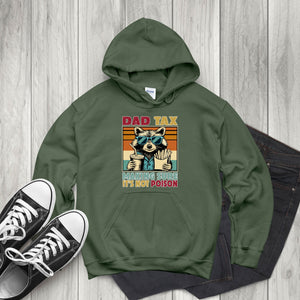 A green hoodie with a graphic design of a raccoon wearing sunglasses and holding a sign that says "DAD TAX MAKING SURE IT'S NOT POISON". The hoodie is placed on a wooden surface, and there are a pair of black and white sneakers visible in the background.
