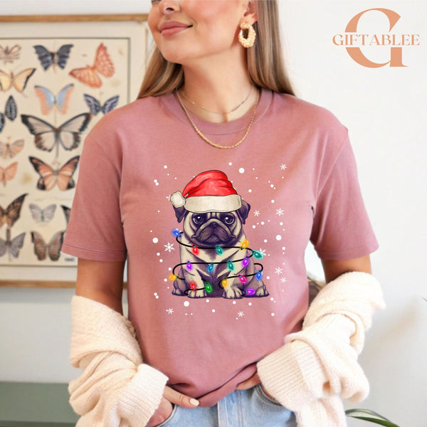 A woman wearing a pink t-shirt with a graphic of a pug dog wearing a Santa hat and surrounded by Christmas lights.
