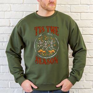 A man wearing a green sweater with a graphic design featuring a skeleton and pumpkin motif, along with the text "Tis the season" printed on it.