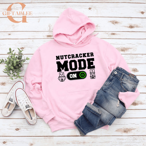 A pink hoodie with the text "NUTCRACKER MODE ON" printed on it, along with a pair of jeans and white sneakers, all laid out on a wooden surface.