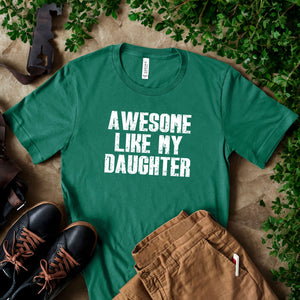 A green t-shirt with the text "AWESOME LIKE MY DAUGHTER" printed on it, along with a pair of brown pants and black shoes, all laid out on a surface with some greenery.