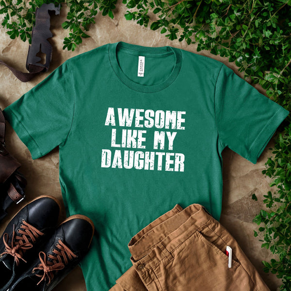 A green t-shirt with the text "AWESOME LIKE MY DAUGHTER" printed on it, along with a pair of brown pants and black shoes, all laid out on a surface with some greenery.