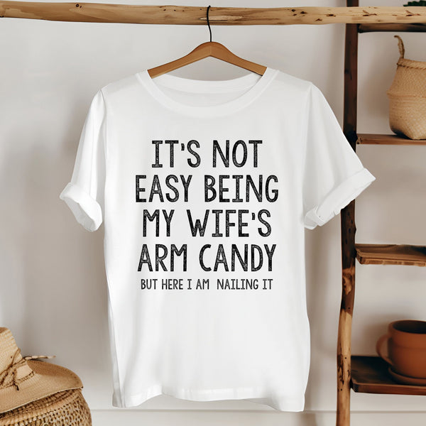 A white t-shirt with a humorous text printed on it, hanging on a wooden hanger.