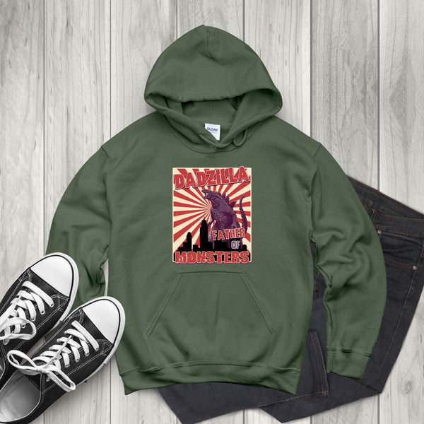 A green hoodie with a graphic design featuring a large image of a dinosaur and the text "DADZILA, FATHER OF MONSTERS" printed on it, along with a red and white sunburst design. The hoodie is placed on a wooden surface, and there are a pair of black and white sneakers visible in the image.