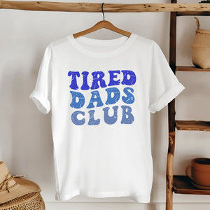 A white t-shirt with the text "TIRED DAD'S CLUB" printed on it, hanging on a wooden hanger.