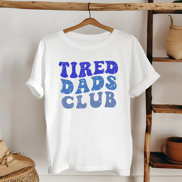 A white t-shirt with the text "TIRED DAD'S CLUB" printed on it, hanging on a wooden hanger.