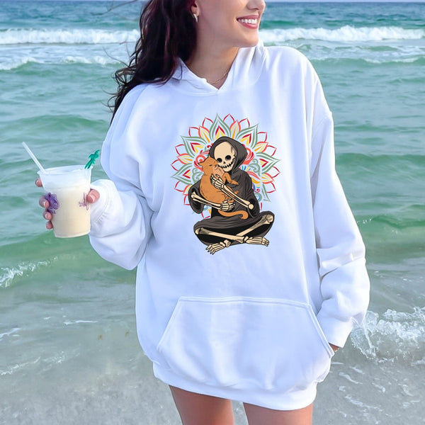 A woman wearing a white hoodie with a graphic of a skeleton holding a cat is standing on a beach, holding a drink in her hand.