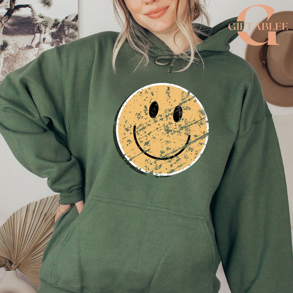 A woman wearing a green hoodie with a distressed yellow smiley face design.