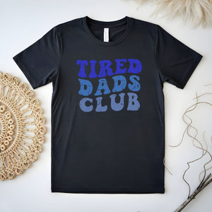 A black t-shirt with the text "TIRED DAD'S CLUB" printed on it, along with a beige woven mat and some twigs in the background.