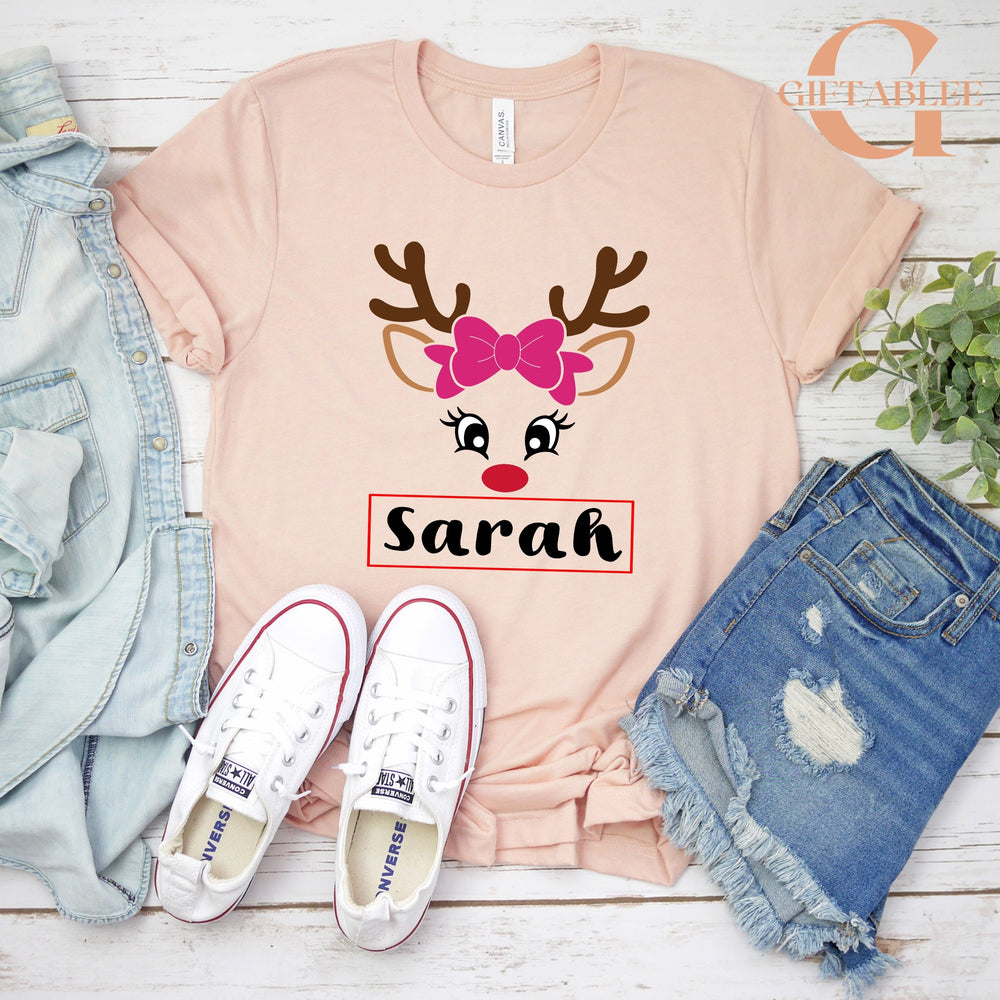 A pink t-shirt with a reindeer face design and the name "Sara" printed on it, along with a pair of white sneakers and a pair of blue denim shorts.